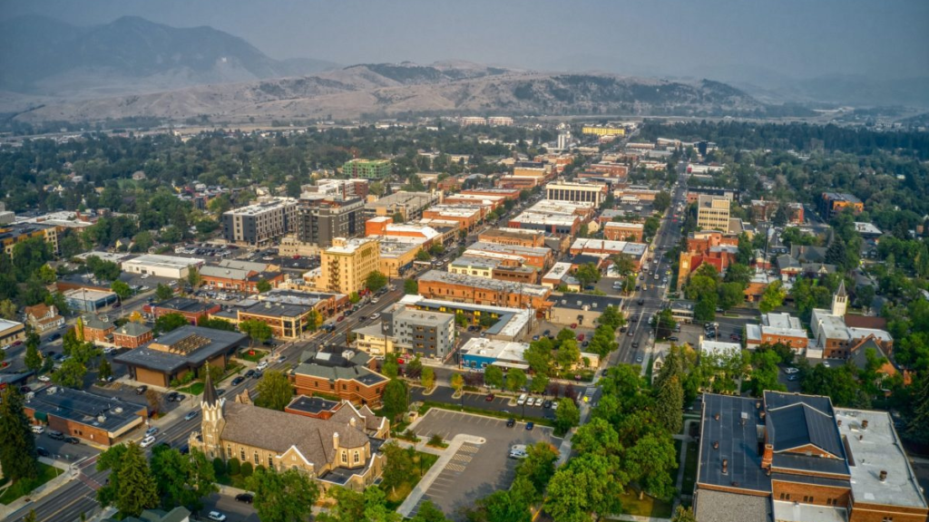 Aerial view of Bozeman, Montana, showcasing its urban landscape with a mix of modern and historic buildings, tree-lined streets, and a backdrop of rolling mountains shrouded in a light haze.