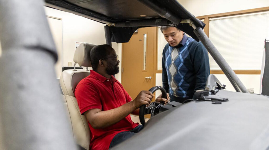 A man sitting in an autonomous vehicle holding the steering wheel while another man looks on