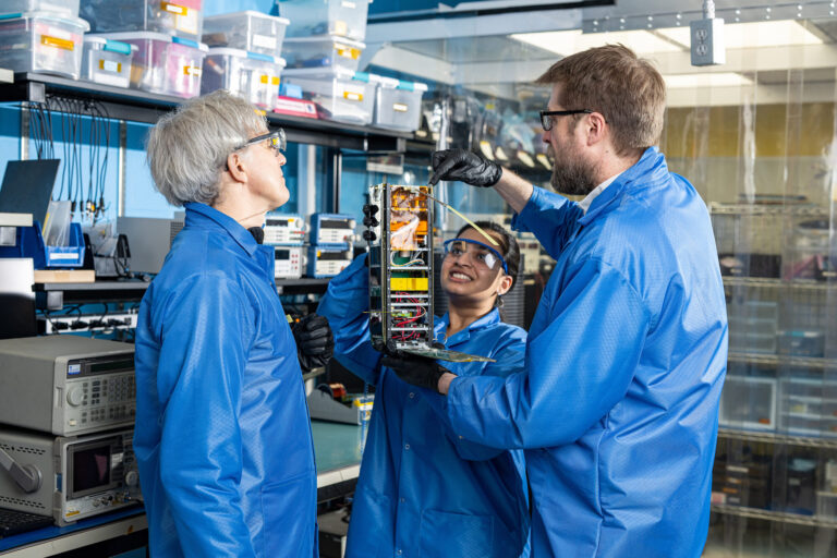 Three researchers wearing blue lab coats and black gloves are talking in a group while examining a small, boxy satellite held by the researcher in the center. The satellite is about a foot tall and four inches wide and deep.
