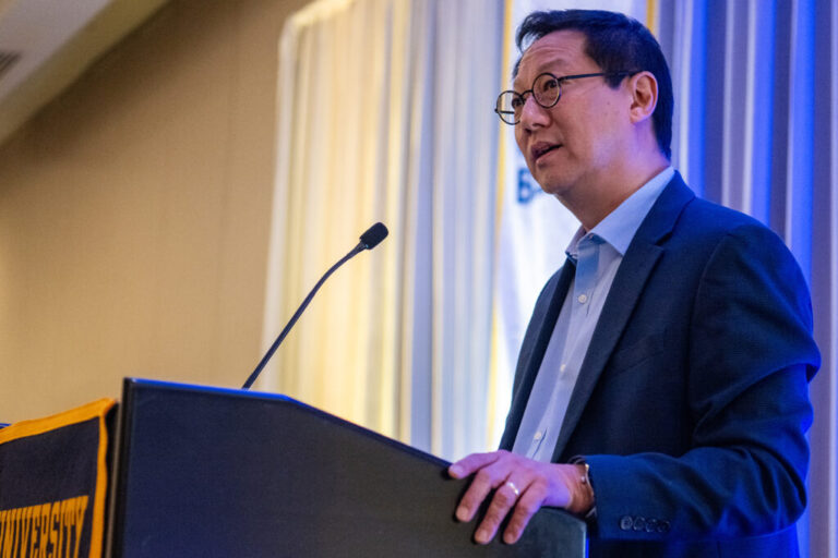 A photo of a man standing and talking at a microphone