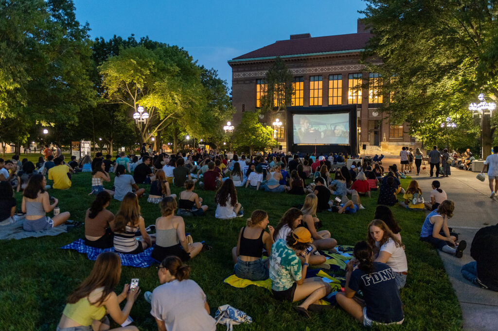 Students sit on grass watching a movie