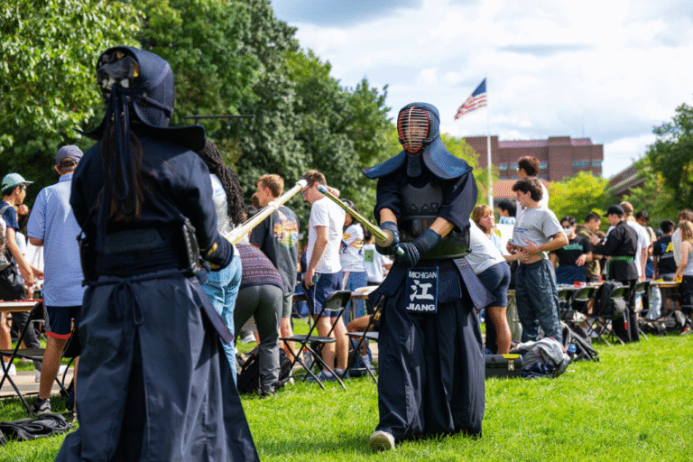 Two people cloaked in black face each other with swords