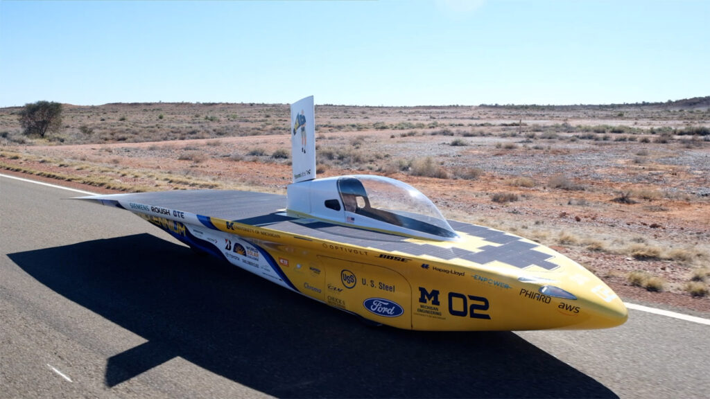 A torpedo-shaped, maize and blue car drives along a paved highway cutting through scrubby red-orange desert sand