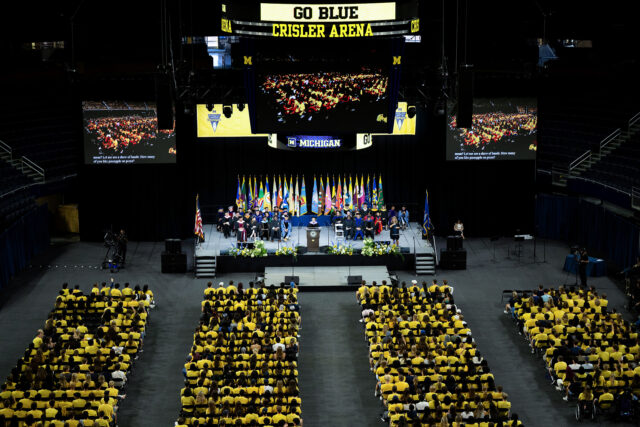 An overhead view of a gathering of people, with seating in four sections and others sitting on a stage