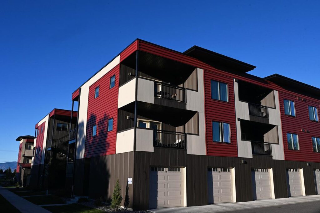 Bozemans WARD Initiative Water and Rent at the Ballots Forefront - Kitchen Table News Modern three-story apartment building with red, beige, and dark brown siding, balconies, and garages on the ground level, under a clear blue sky.