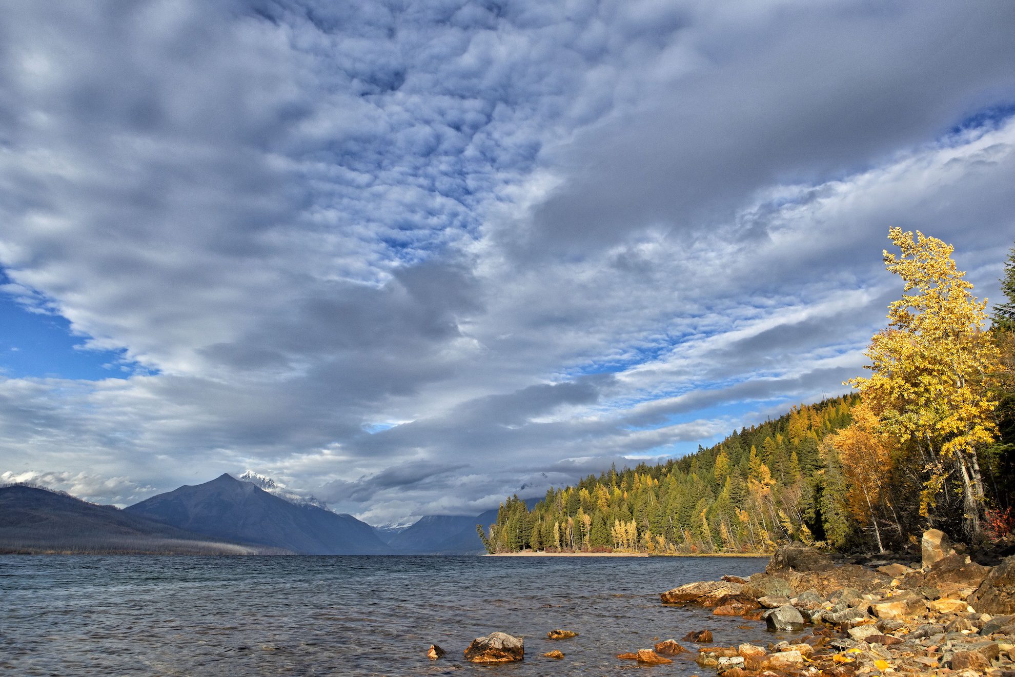 Government Shutdown Impacts Tourism at Glacier National Park Economy Hurts - Kitchen Table News Lake McDonald with a rocky shoreline, autumn trees, and distant snow-capped mountains