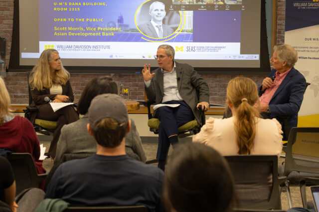 Three people, one of them speaking, facing an audience seated in chairs