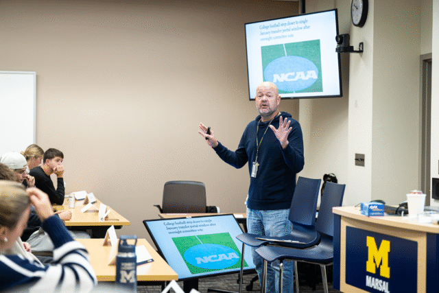 A man stands before a class of students and speaks