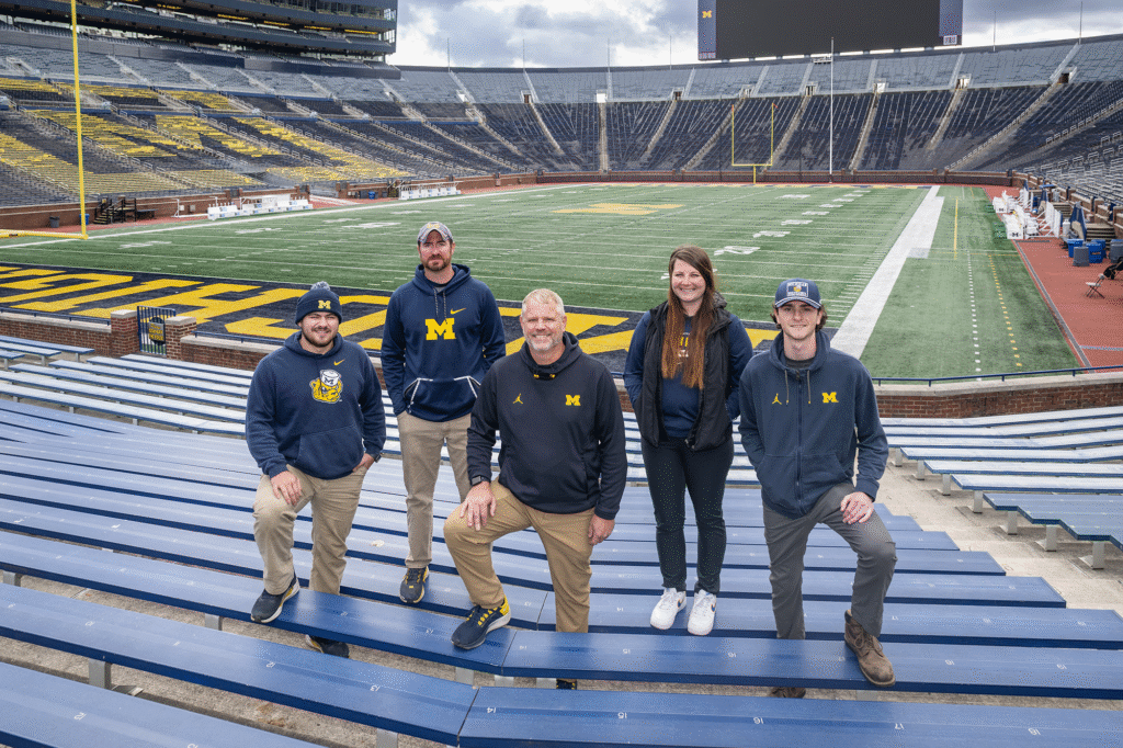 Five people stand in the grandstands of Michigan Stadium with the field behind them