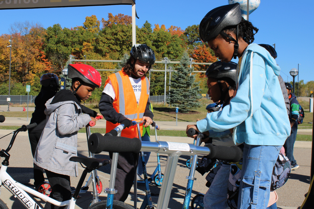 A man wearing a bike helmet oversees children wearing bike helmets while they pick out scooters to ride