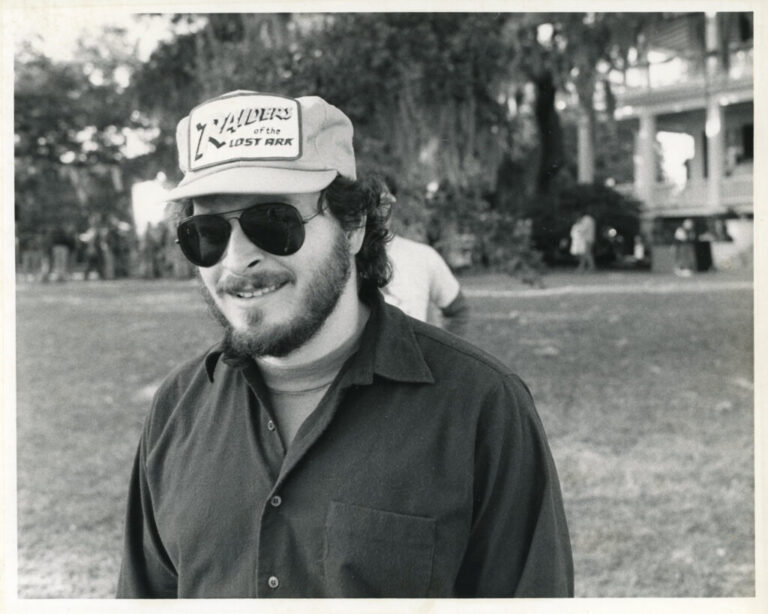A man in sunglasses and wearing a ballcap smiles on the set of a movie