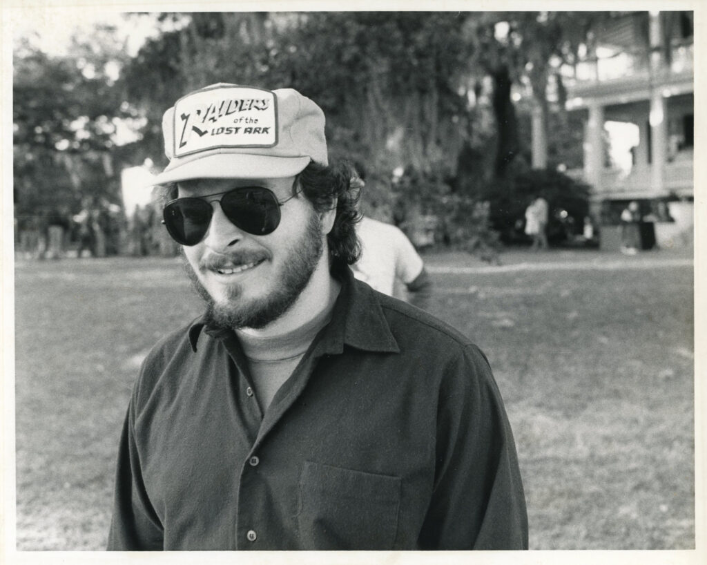 A man in sunglasses and wearing a ballcap smiles on the set of a movie