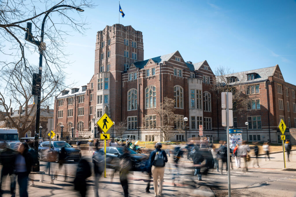 People are blurred walking across the street on a sunny day with a large building in focus in the background