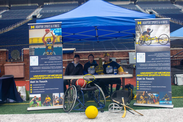 Three people sit behind a table under a tent surrounded by adaptive sports equipment