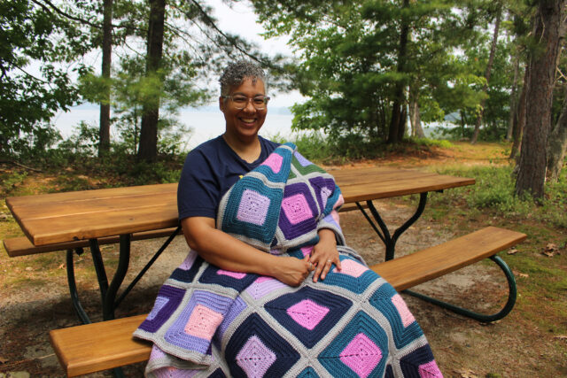 A woman sitting at a picnic table with a lake and woods behind her and a crocheted afghan on her lap