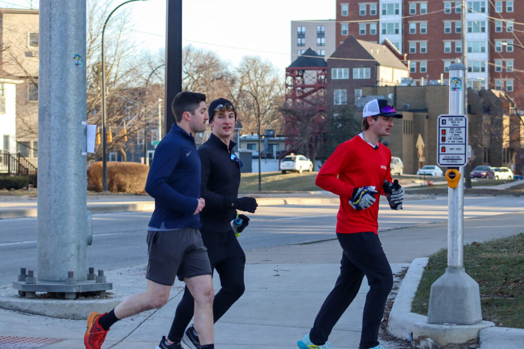 Three men run on a sidewalk on a chilly but sunny day