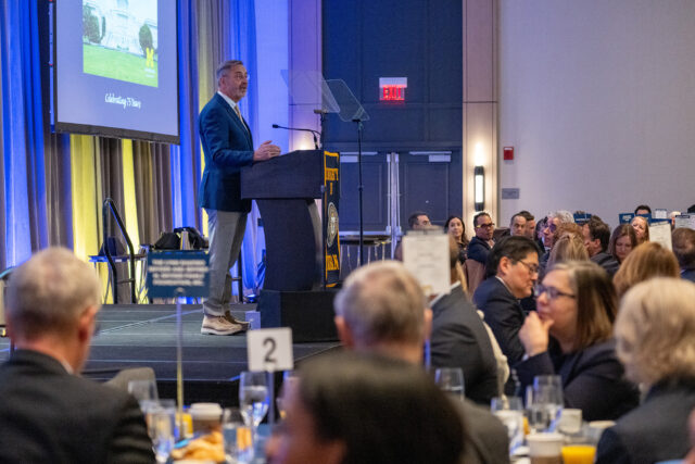 A man speaks at a podium on a stage in front of a large crowd seated at tables