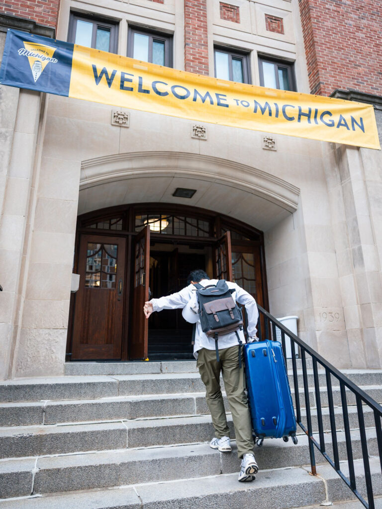 A person carries a suitcase up concrete stairs into a building under a banner that says "Welcome to Michigan"