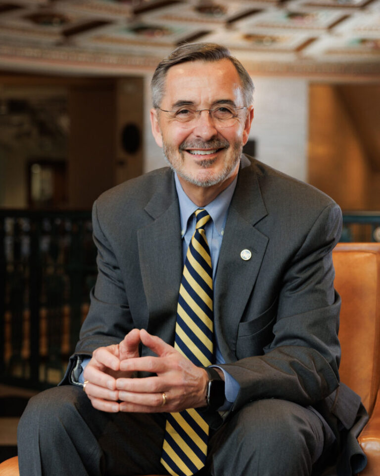Man positing wearing a gray suit, blue shirt, maize and blue tie, and glasses.