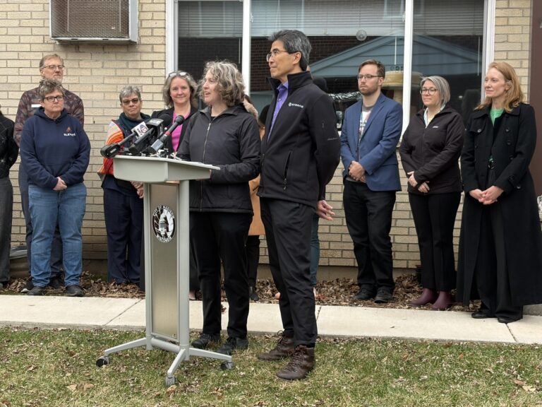 Group of people standing behind a podium with microphones outside a brick building, a city seal visible on the podium.