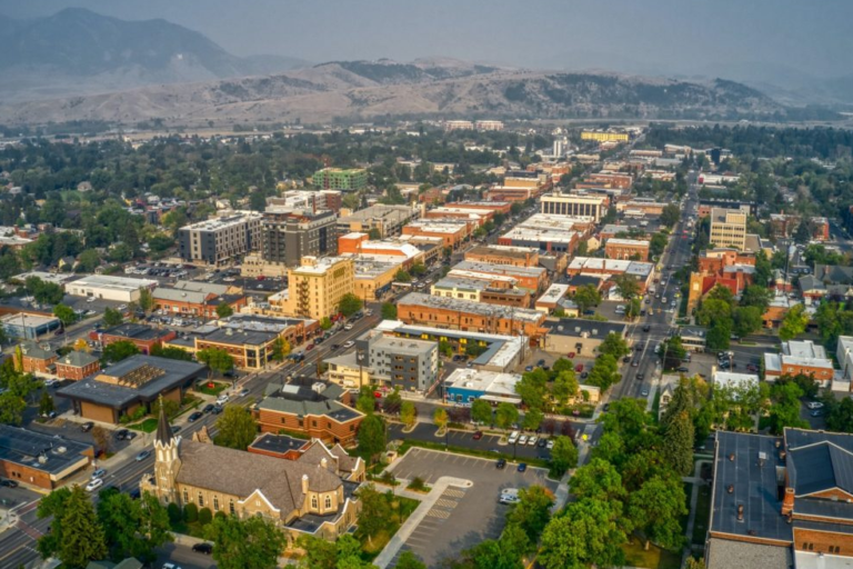 Aerial view of Bozeman, Montana, showcasing its urban landscape with a mix of modern and historic buildings, tree-lined streets, and a backdrop of rolling mountains shrouded in a light haze.