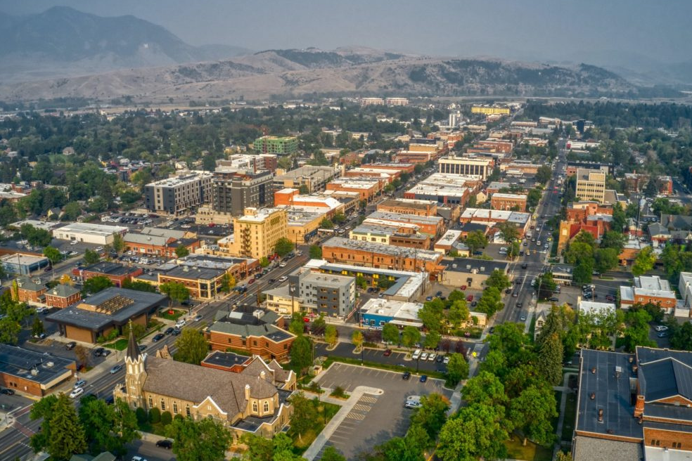 Aerial view of Bozeman, Montana, showcasing its urban landscape with a mix of modern and historic buildings, tree-lined streets, and a backdrop of rolling mountains shrouded in a light haze.