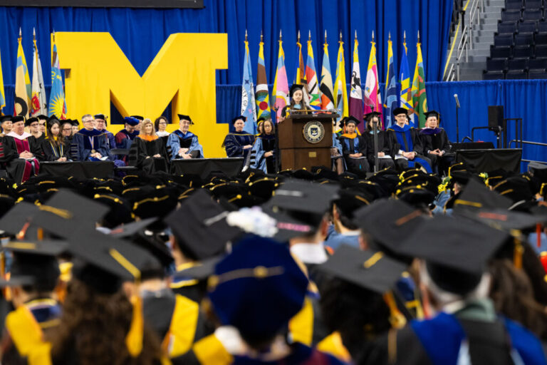 college leaders sitting on stage in graduation regalia listening to a student speaker talk at a podium