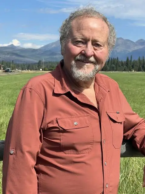 Older man with gray hair and beard, wearing a rust-colored shirt, standing in a grassy field with mountains in the background and a wooden fence nearby.