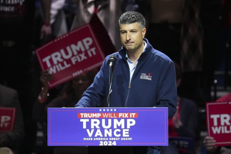 Male speaker at a political rally, standing behind a podium with Trump-themed signage visible in the crowd.