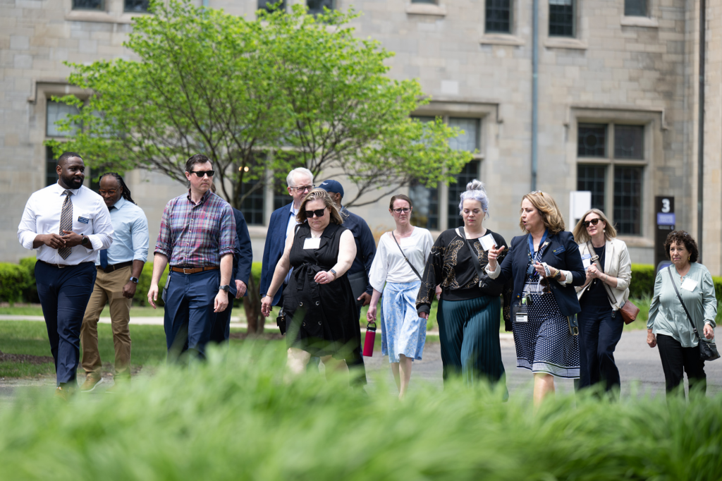 Several people walk outside, some wearing sunglasses, with a large building in the background and blurred grass in the foreground