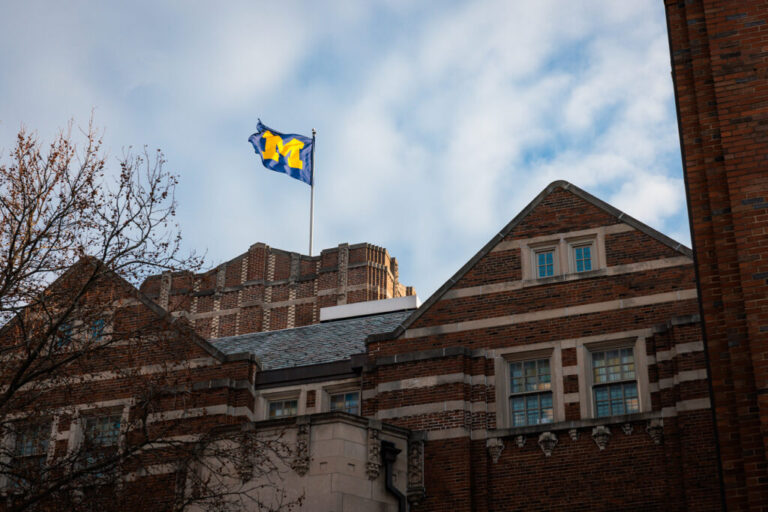 A U-M flag flies atop a building with blue sky and white clouds