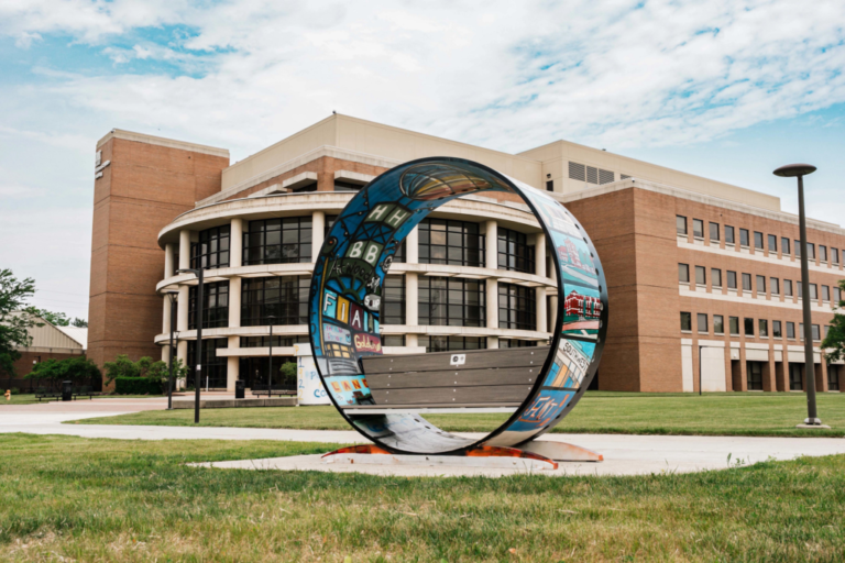 A circular piece of art stands outside a large brick building