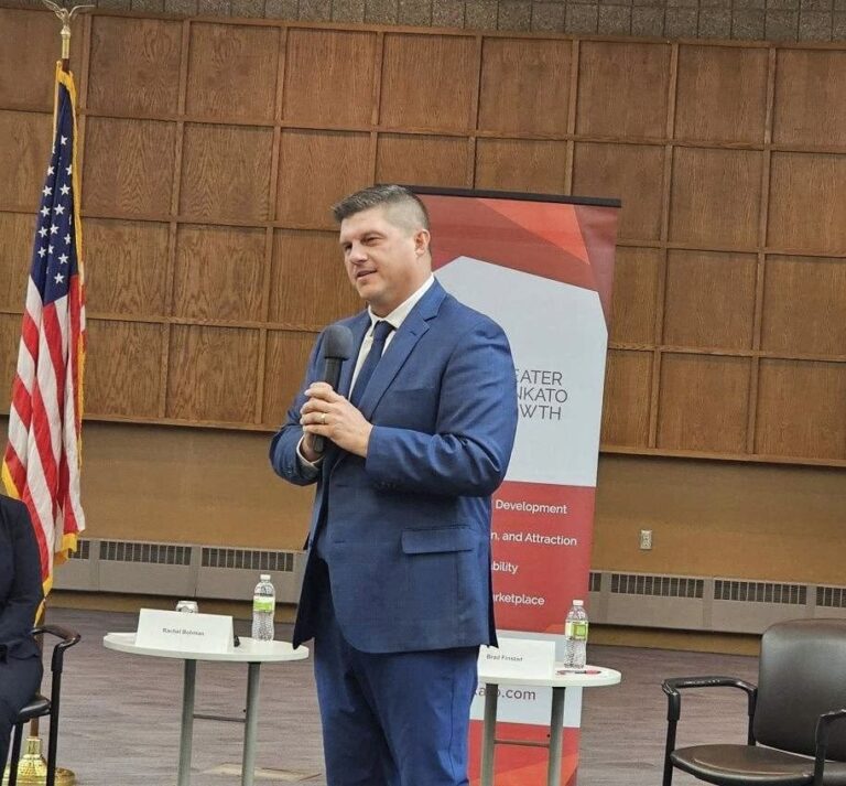 Man in a blue suit speaks into a handheld microphone at a conference, with an American flag and a banner behind him.